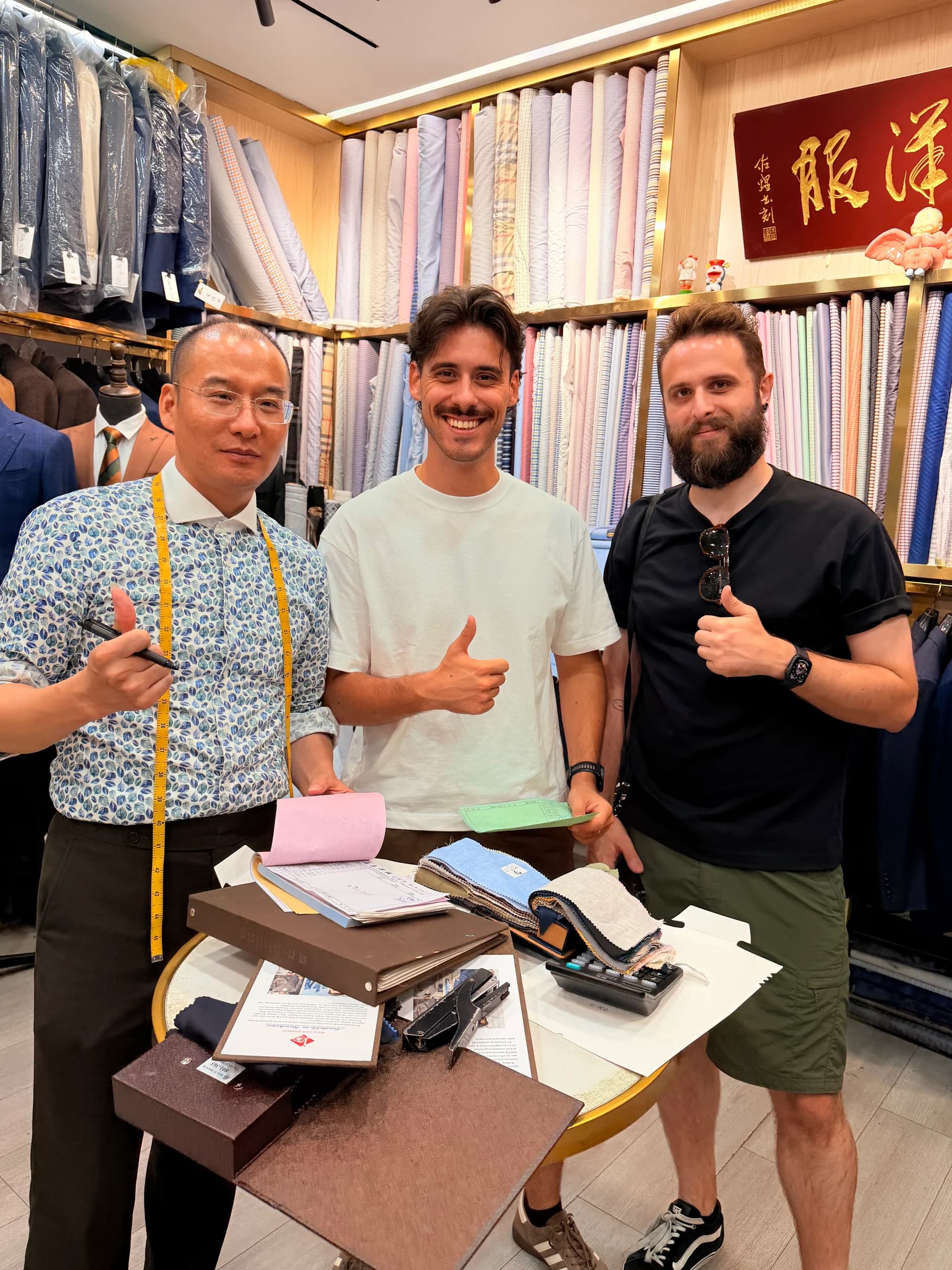 Two international visitors reviewing fabric swatches with a Shanghai fabric market tailor