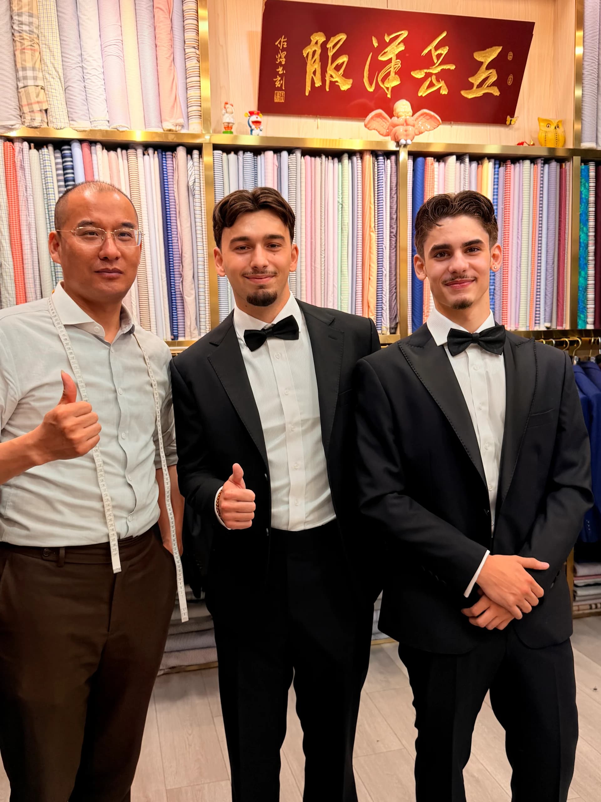 Two clients in finished black tuxedos standing with a Shanghai fabric market tailor