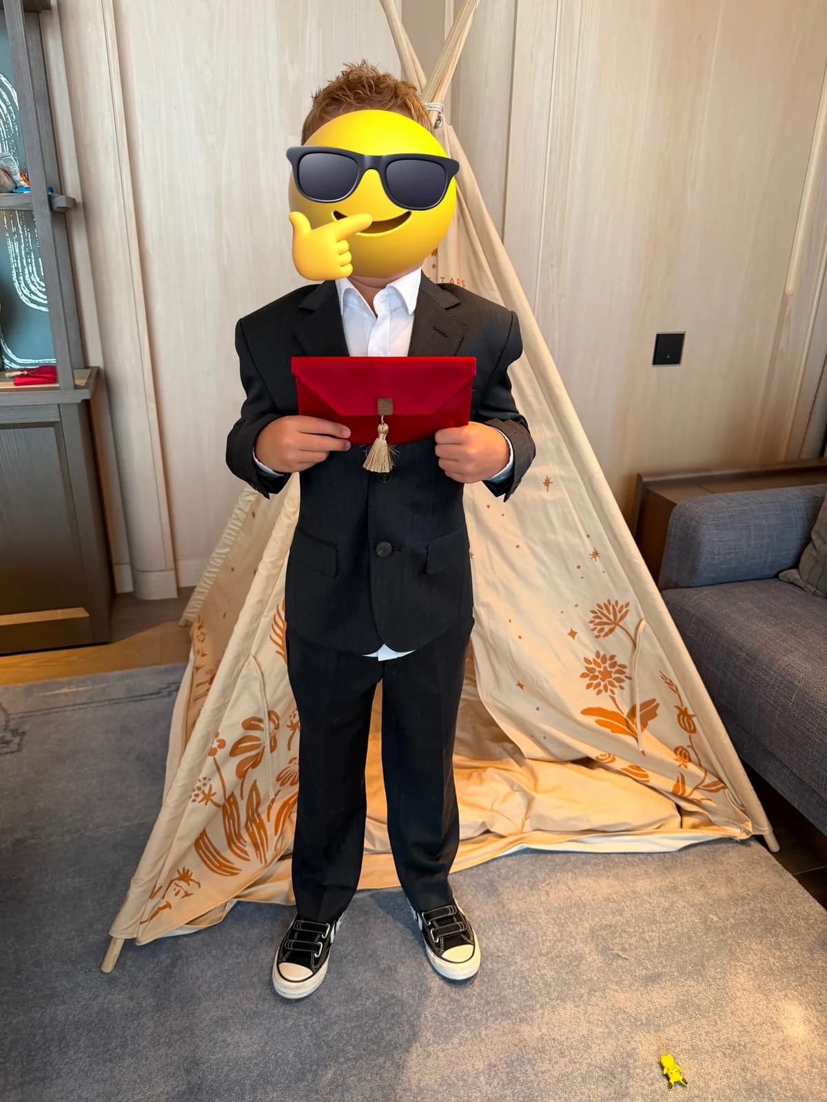 Full-length view of a child in a dark suit, white shirt and trainers, holding a red pouch